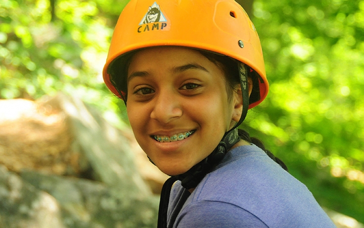 A young person wearing a helmet smiles at the camera. There is blurred greenery in the background. 
